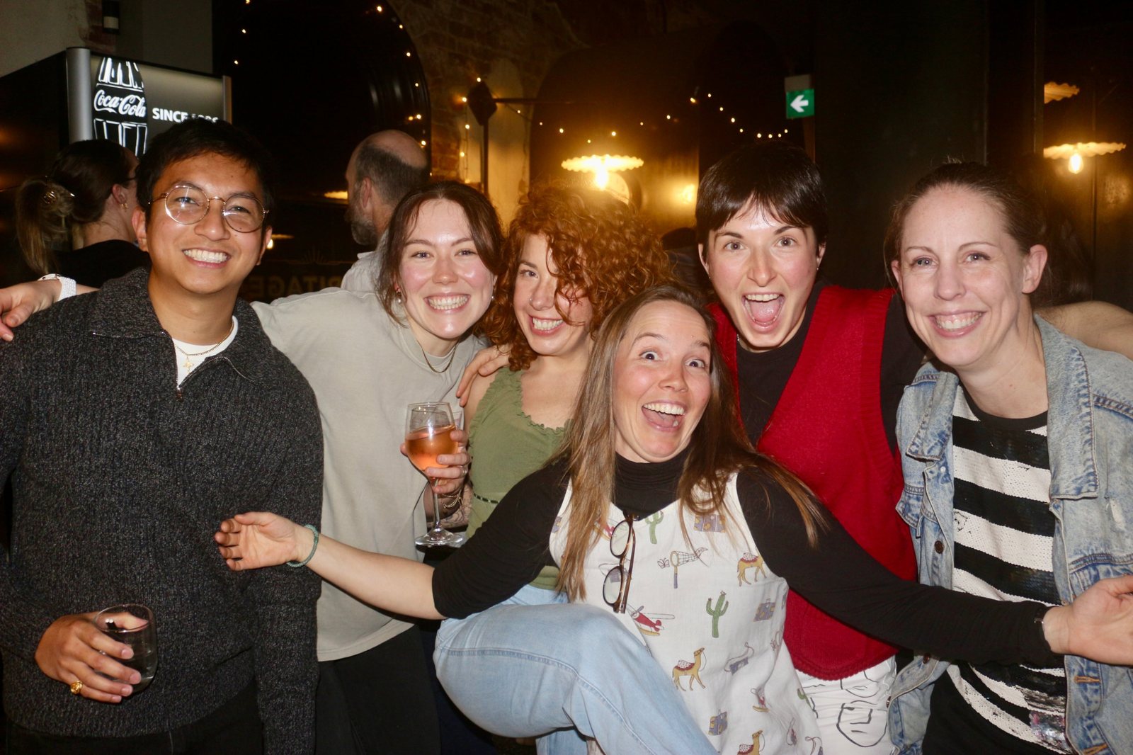 Six friends with drinks, arms around each other at the bar